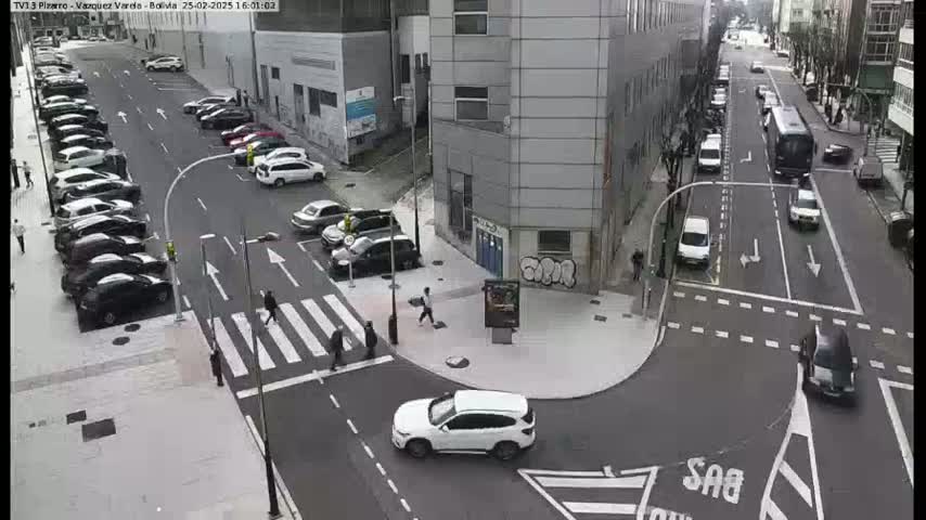A city street intersection with parked cars, pedestrians crossing, and several vehicles moving along the roadways under overcast skies.