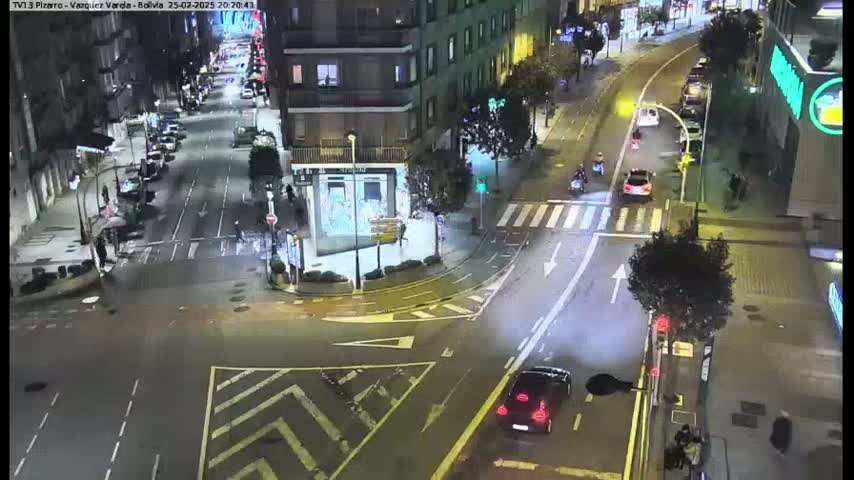 A nighttime aerial view of a city intersection with several vehicles and pedestrians, under clear skies.