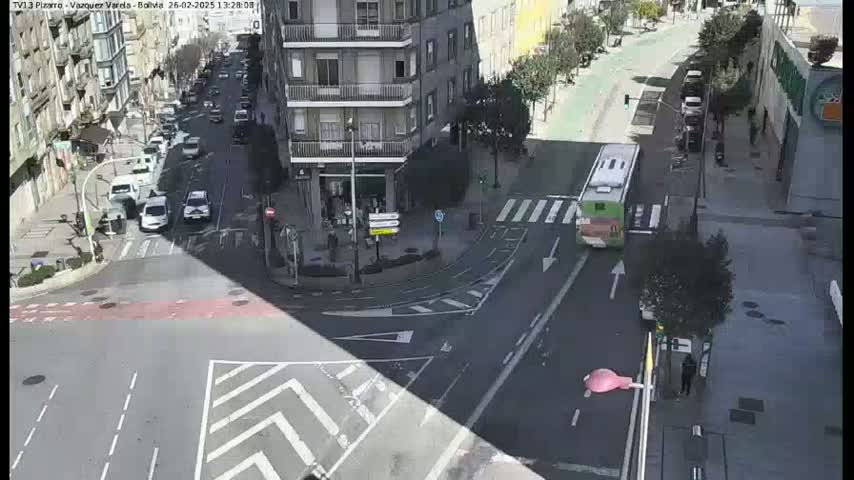 A sunny day reveals a city intersection with several cars and a bus, surrounded by buildings and pedestrian walkways.