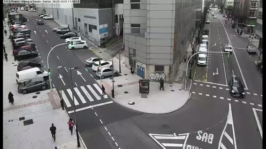 A street intersection with parked cars along the sides and several cars driving through, on a partly cloudy day.