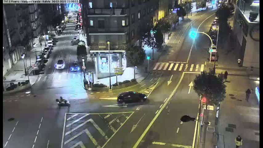 A nighttime street scene shows several vehicles at an intersection, with a few pedestrians and a motorcycle visible under wet conditions.