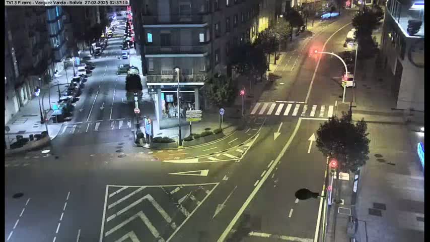 A nighttime, high-angle view of a city street intersection with cars parked along the sides of the road and a few vehicles driving.