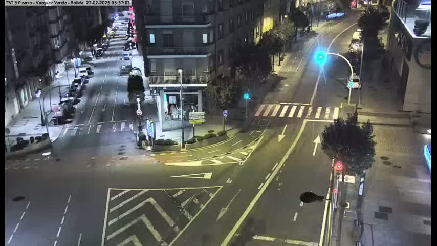 A nighttime aerial view of a city intersection with several cars parked along the sides of the street and a few cars traveling through the intersection under wet conditions.