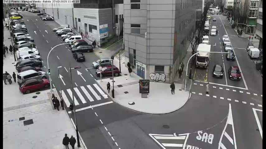 A city street intersection with parked and moving cars, pedestrians, and buildings, on an overcast day.