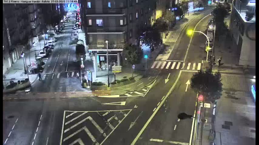 A wet, nighttime street intersection with cars parked along the sides of the road and a few pedestrians walking.