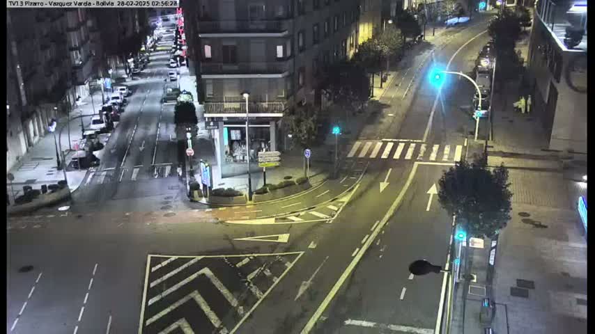 A nighttime, overhead view of a city intersection with several cars parked along the sides of the streets and a few cars driving.