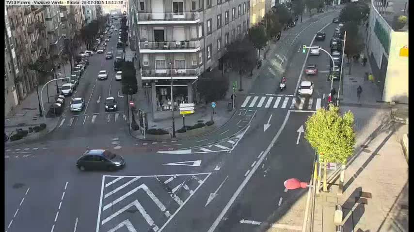 A high-angle, sunny-day view of a city intersection with several cars driving on the roads, pedestrians on the sidewalks, and buildings lining the streets.