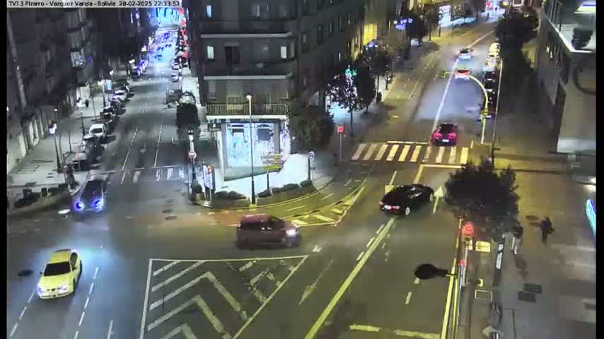 A nighttime street intersection shows several cars at a crossroads, with pedestrians and streetlights visible under partly cloudy skies.