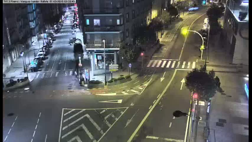 A nighttime, high-angle view of a city intersection with several cars parked along the sides of the streets and a few cars driving, showing a street with crosswalks and traffic signals.