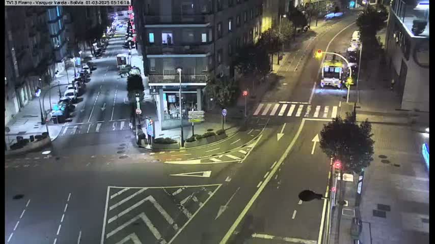 A nighttime high-angle view of a city street intersection with several parked cars and a single bus driving through.