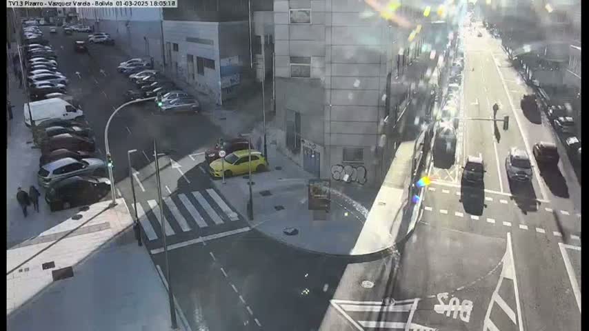 A sunny day shows a city intersection with parked and moving cars, pedestrians, and buildings.