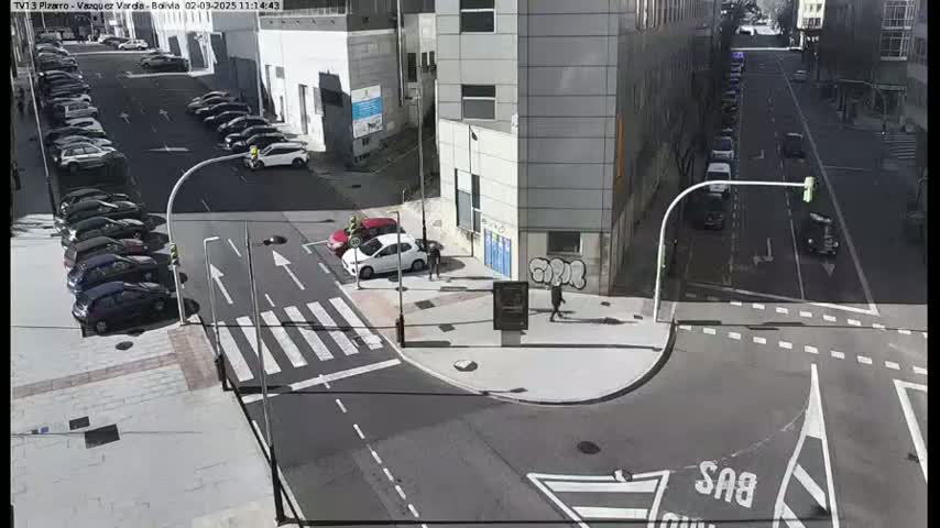 A sunny city street intersection shows several parked cars along the sides of the street and a few cars driving through the intersection.