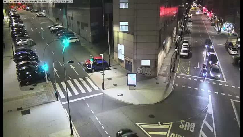 A nighttime street intersection shows several parked cars along the side of the road and a few moving vehicles in the intersection, with a person on a motorcycle visible in the middle.