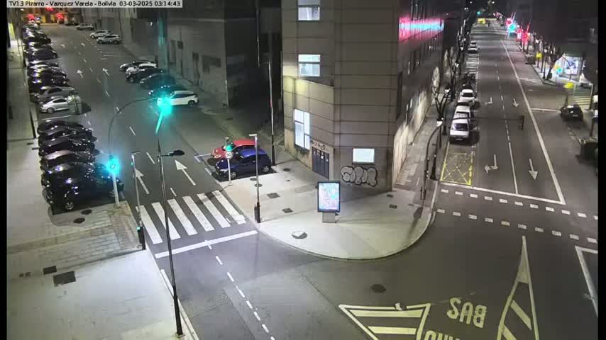 A nighttime street intersection shows several parked cars along the sides of the roads, and a few cars driving down the roads.