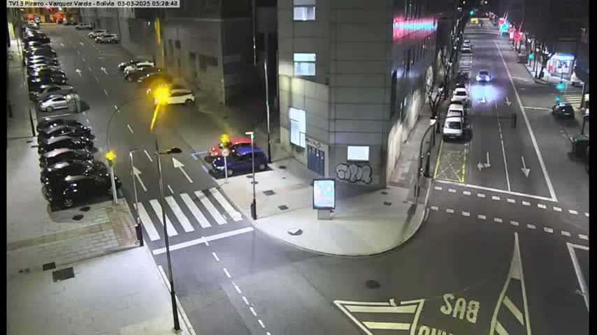 A nighttime street scene shows several cars parked along the sides of a road that intersects with another road, with additional cars driving on both roads.