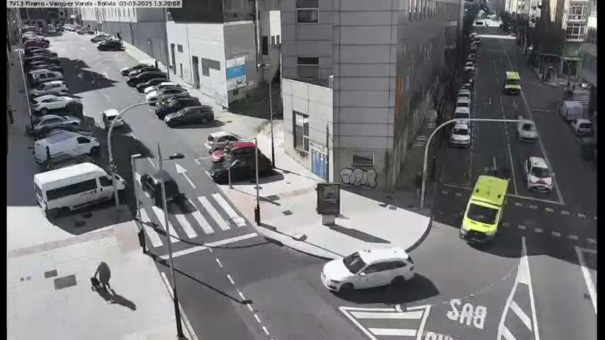 A street scene on a sunny day shows a busy intersection with cars parked along the sides of the street and driving on the road, an ambulance stopped at the intersection, and a pedestrian walking along the sidewalk.