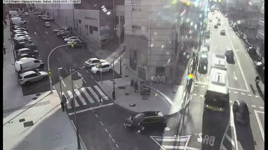 A sunny city street intersection shows parked cars along the sides of the road, and moving vehicles at a crosswalk and further down the road.