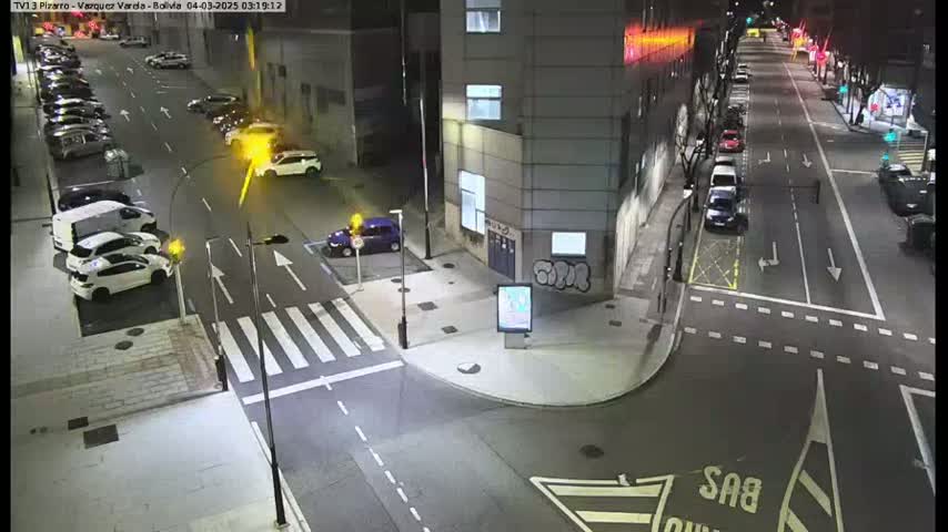A nighttime street scene shows several cars parked along the side of a street and others driving on a multi-lane road that intersects with a crosswalk.