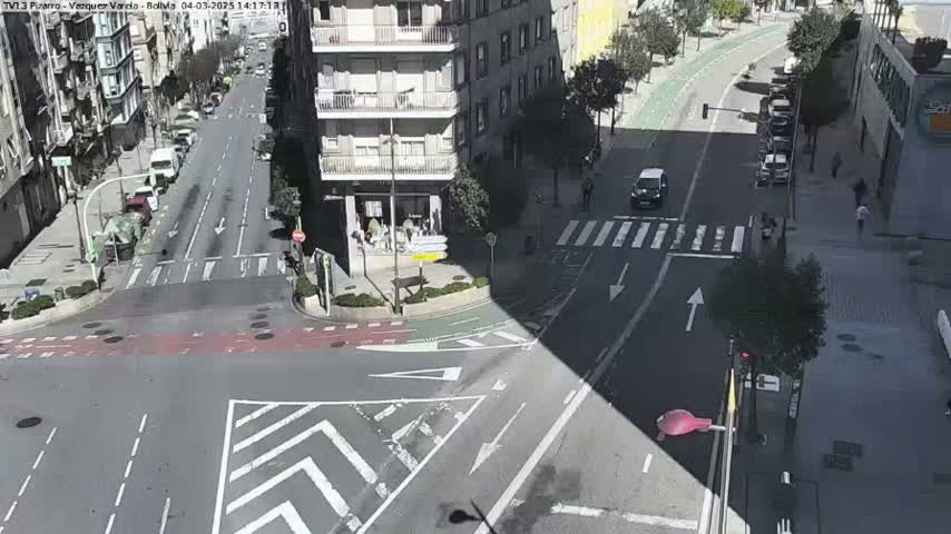 A sunny street intersection in a city shows several cars and pedestrians, with a bike lane visible alongside the road.
