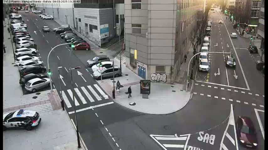 A city intersection with parked cars lining the streets and a few pedestrians walking, under a mostly sunny sky.