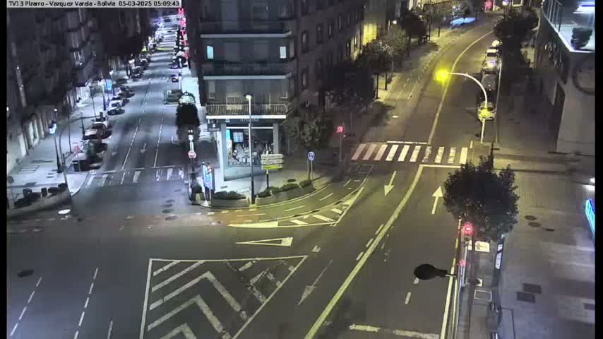A nighttime, street-level view of a city intersection with several cars and buildings visible under artificial lighting.