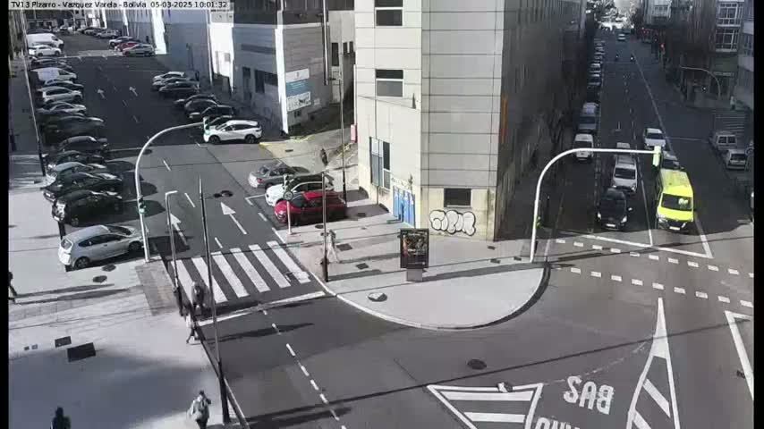 A sunny city intersection shows several cars parked along the street and others driving through the intersection, pedestrians walking on a crosswalk, and buildings in the background.