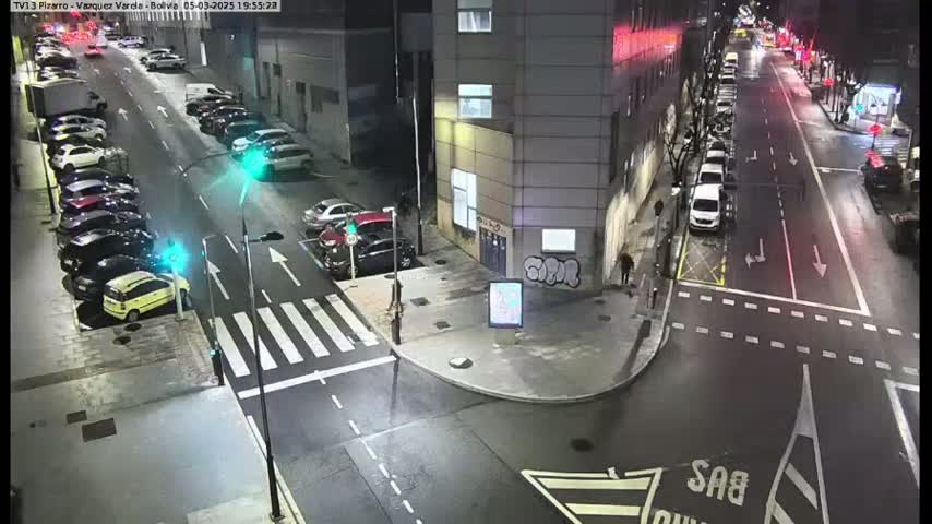 A nighttime street scene shows cars parked along the sides of a street that intersects with another street, and there are traffic lights at the intersection.