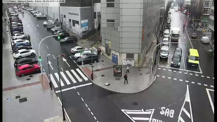 A wet city street intersection shows several cars parked along the sides and a few driving through, with pedestrians near a building.
