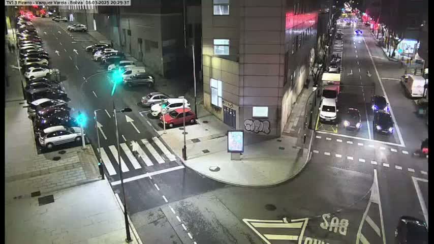 At night, a street intersection shows several parked cars along the side of a road and other cars driving on the road.