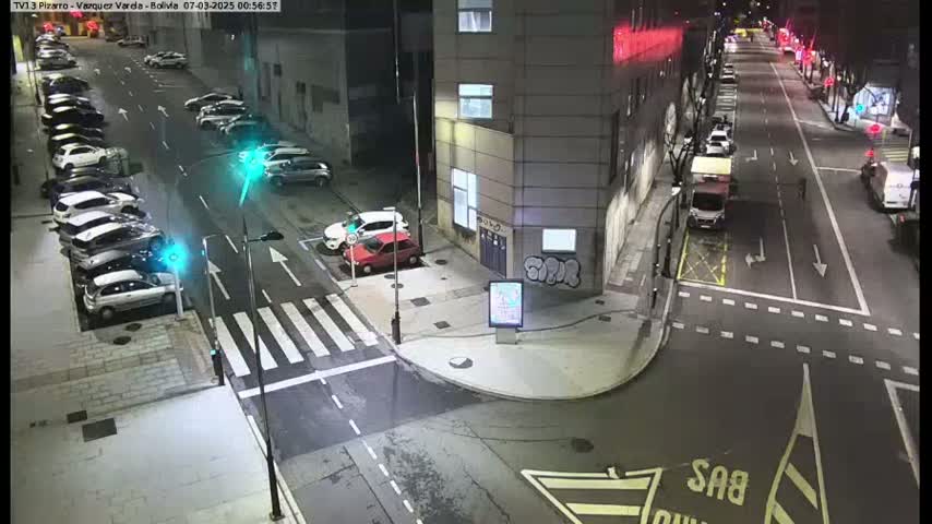 A nighttime street scene shows cars parked along the side of a building and a few driving on the street, with a crosswalk and traffic signals.
