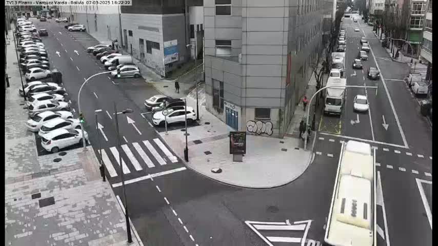 A street intersection with parked cars and moving vehicles, under a cloudy sky.