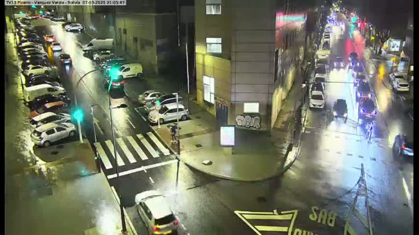 A rain-slicked city street at night shows numerous parked cars and several cars driving on the street, with some pedestrians visible near a crosswalk.