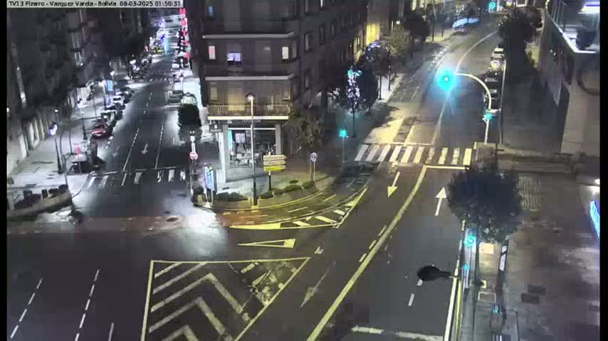 A wet, nighttime street intersection shows several cars parked along the sides of the road and a few moving through the intersection under dim streetlights.