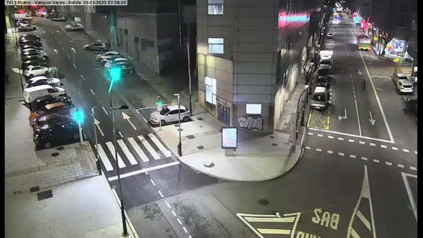 A nighttime street scene shows several cars parked along the sides of a street and a few driving on the roadway, with a crosswalk and traffic lights visible.