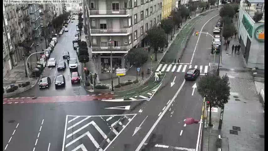 A wet city street intersection shows several cars navigating a crosswalk and bike lane, with pedestrians and buildings in the background.
