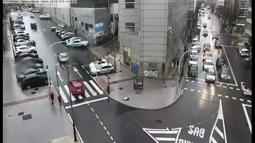 A rainy city intersection shows multiple cars parked and driving on wet roads, with several buildings and pedestrians visible.