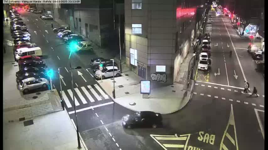 A nighttime street intersection with cars parked along the sides of the road and several vehicles moving through the intersection.