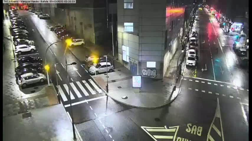 A wet street at night shows several parked cars along the sides and a few cars driving on the road, with a building in the background.