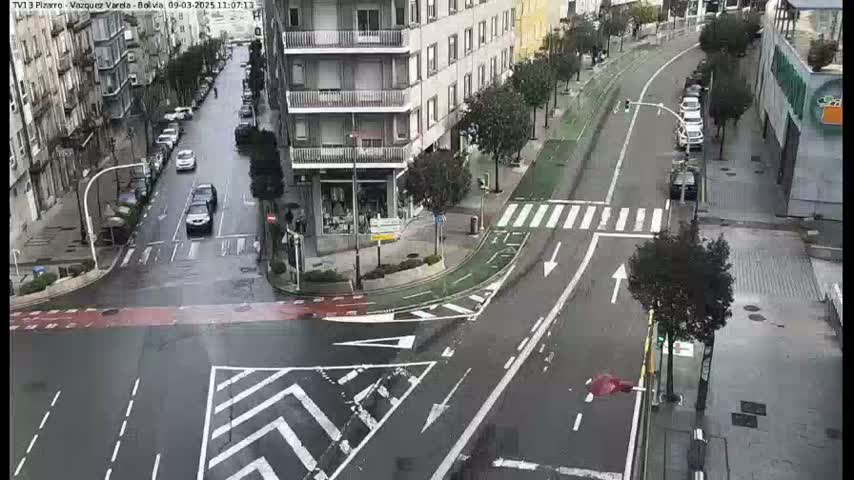 A wet city street intersection with several cars and a bike lane, viewed from above.
