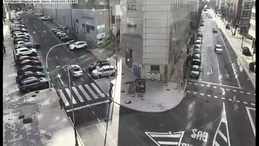 A street intersection with parked cars lining the sides of the road and several cars driving through the intersection on a slightly overcast day.