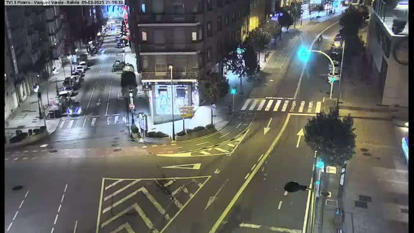 A nighttime, high-angle view of a city intersection with several cars parked along the sides of the streets and a few cars driving through the intersection.