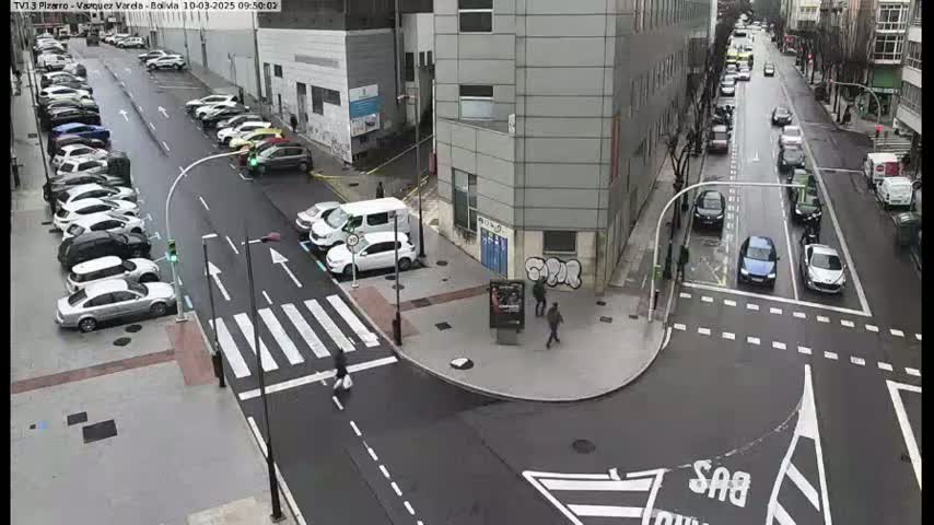 A city street scene with parked cars lining both sides of the road, several cars driving down the street, and a pedestrian crossing, on an overcast day.
