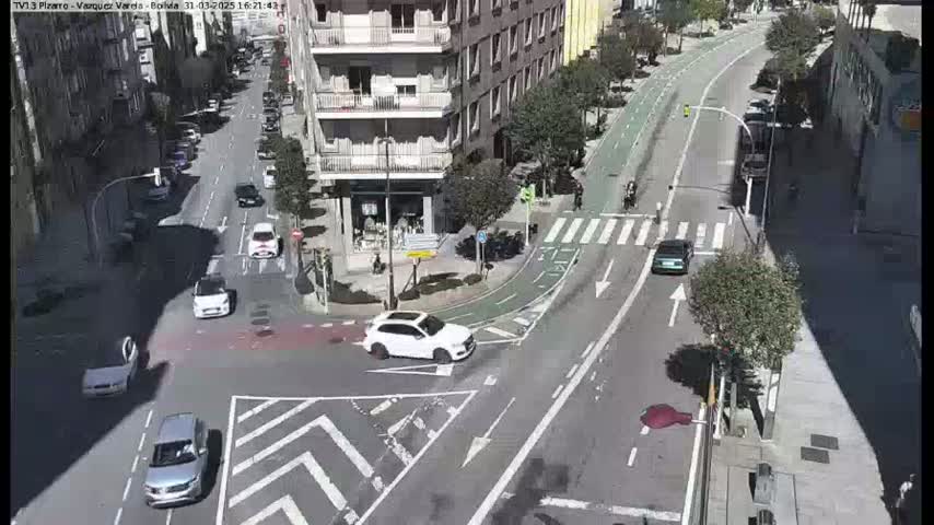 A sunny day shows several cars at a city intersection with a bike lane, near a multi-story building.