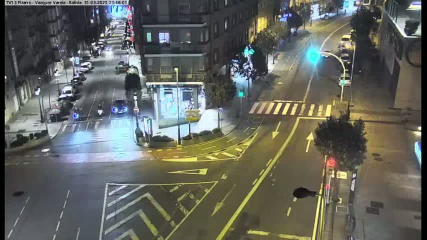 A nighttime, wet street intersection with several cars and a few pedestrians is seen from an elevated viewpoint.