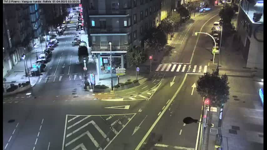 A nighttime, high-angle view of a city street intersection with parked cars along the sides and a few vehicles on the road.