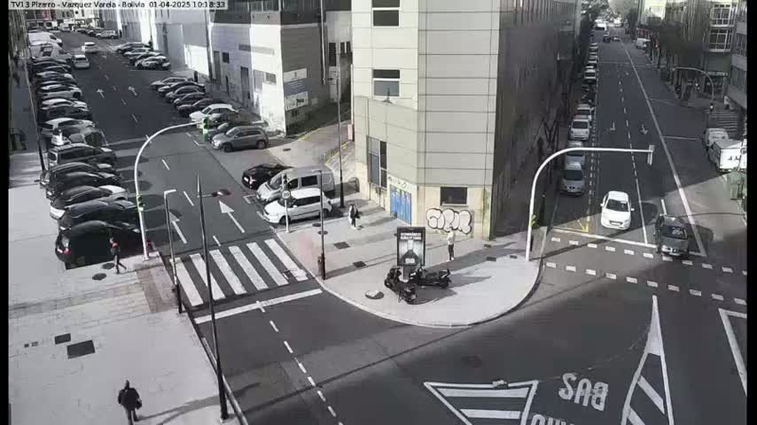 A sunny city street intersection shows several cars parked along the sides and a few driving through, with pedestrians walking near a crosswalk.