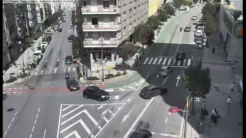 A sunny city intersection shows several cars at a crosswalk and several pedestrians near a building.