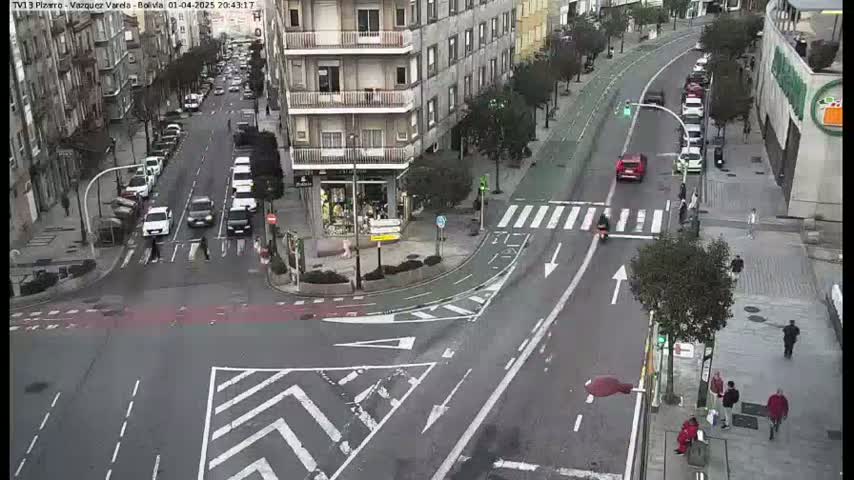 A high-angle, daytime view of a city intersection with several cars and pedestrians, and a mostly clear sky.