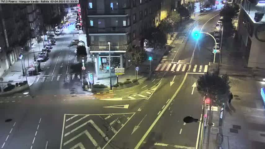 A nighttime, wet street intersection shows several cars parked along the sides of the roads and a few cars driving through.