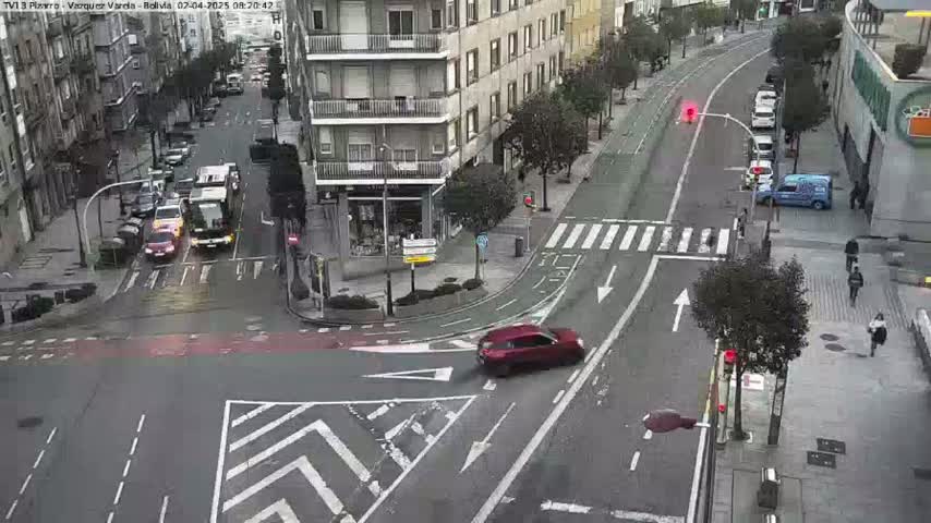 A multi-lane road intersection with several cars and pedestrians, under an overcast sky.